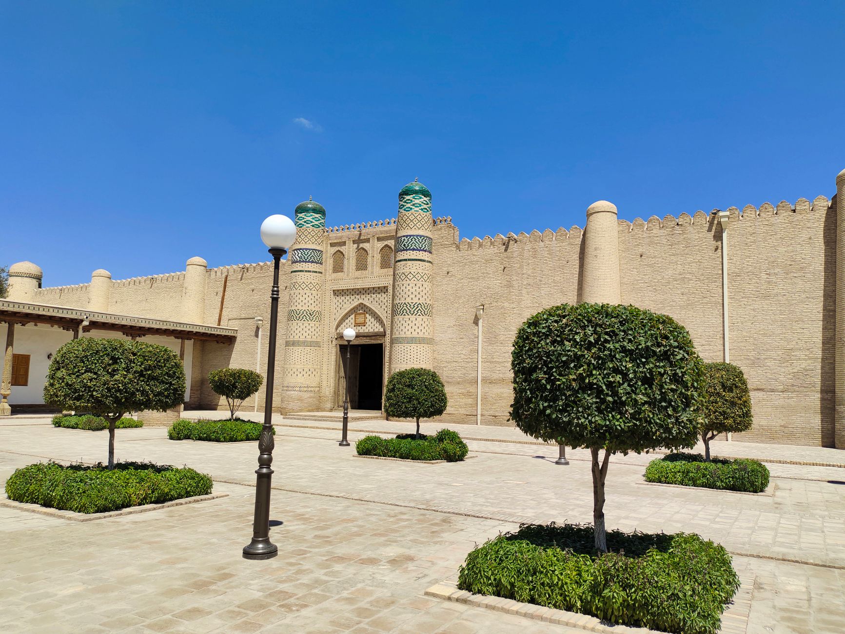 A historic fortress wall in Bukhara, Uzbekistan, lit by bright daylight, showcasing traditional architecture and manicured greenery