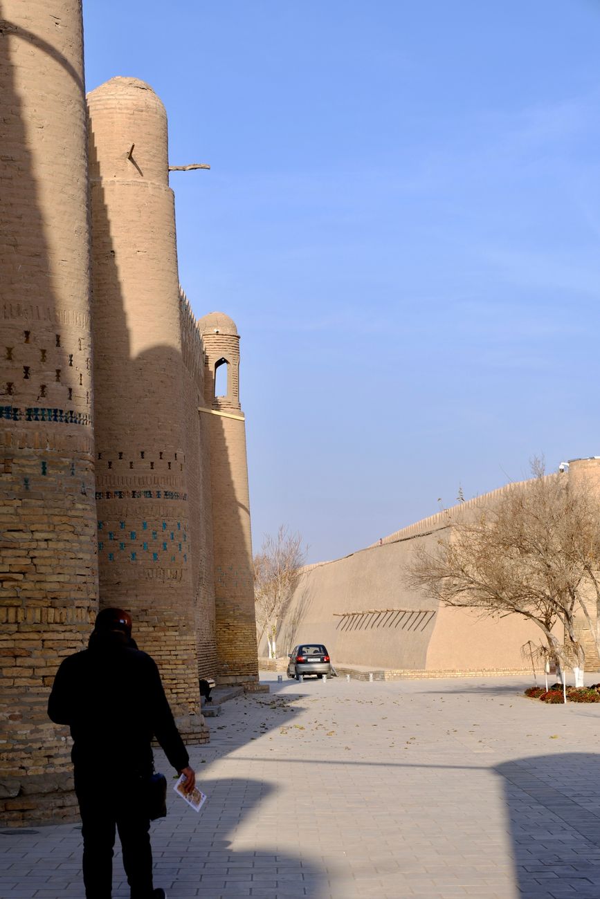 A person walks near the historic mudbrick walls of Khiva, Uzbekistan, bathed in sunlight · History, Uzbekistan